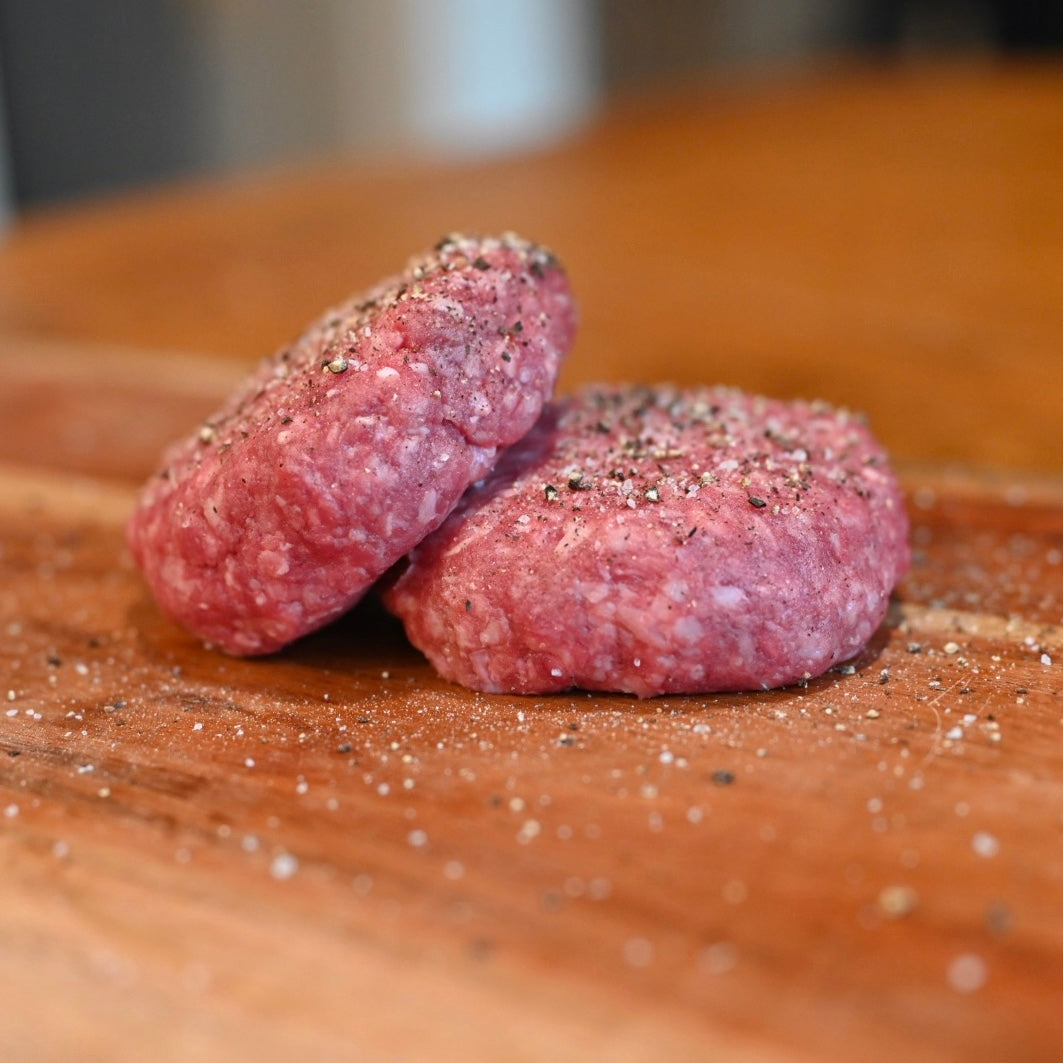 Two raw meat patties on a wooden surface with seasoning.