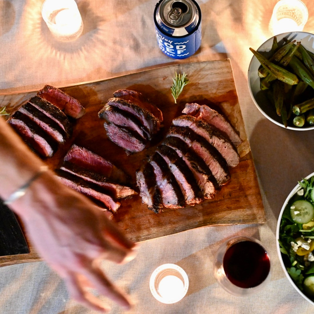 Sliced steak on a wooden board with a can of beer and side dishes on a table.